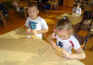 Dwoje dzieci siedzi przy stole, w rękach trzymają gumki recepturki, które zakładają na kartonowe małe pudełko.